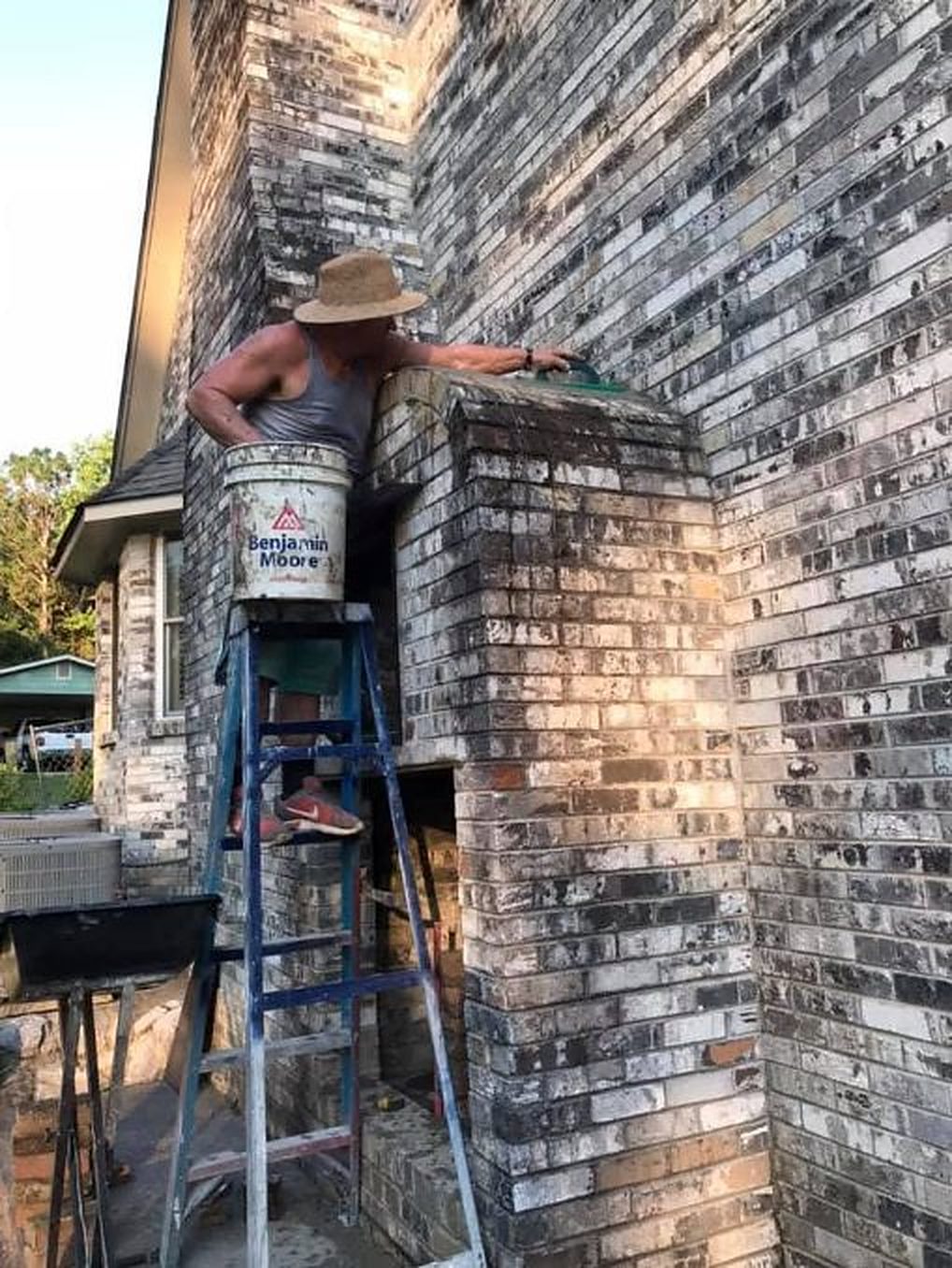A mason on a ladder working on a brick chimney, with his mortar bucket hung on the ladder