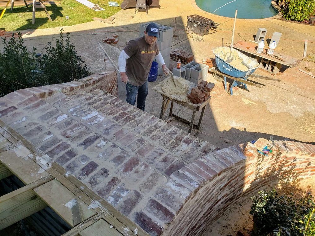 A mason at work beside a curved brick wall, with the mortar board set up and a pool behind