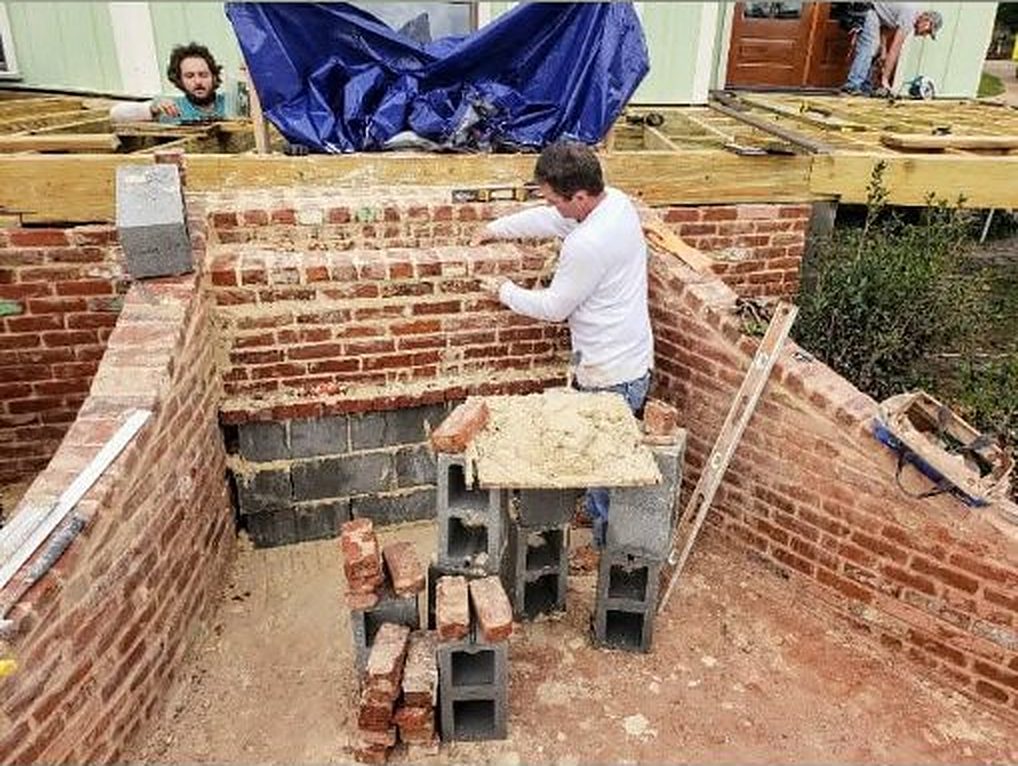 Masons laying old brick walls in a dug out space beside a house, under new deck framing
